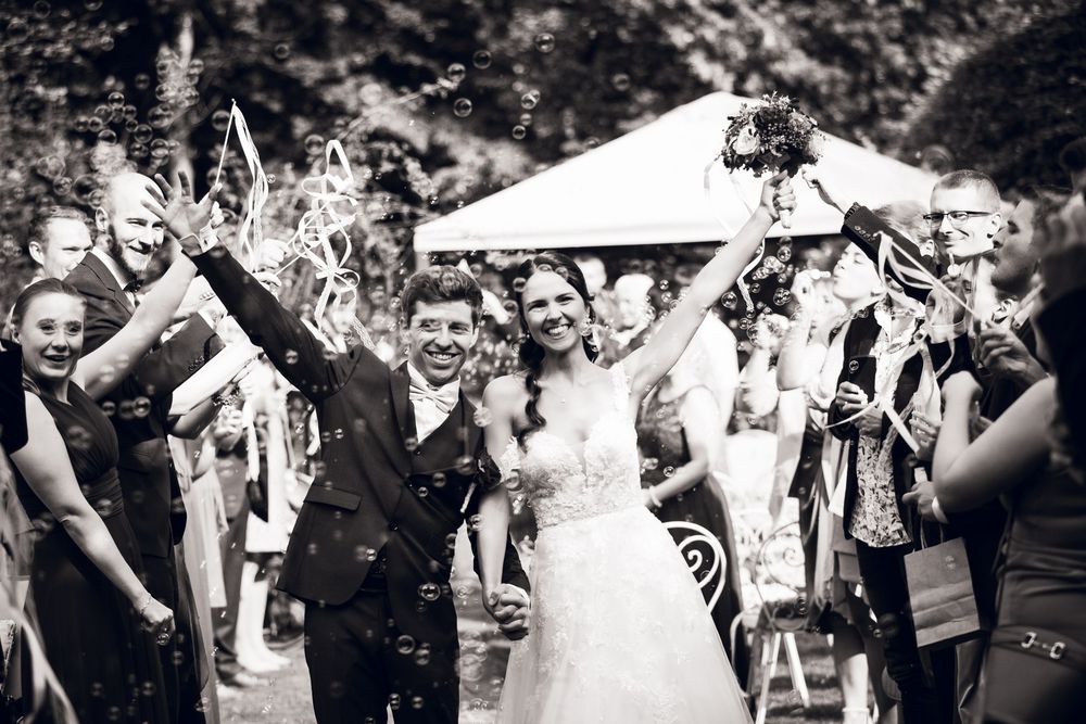 Joyful bride and groom walk through cheering guests blowing bubbles at outdoor wedding reception.