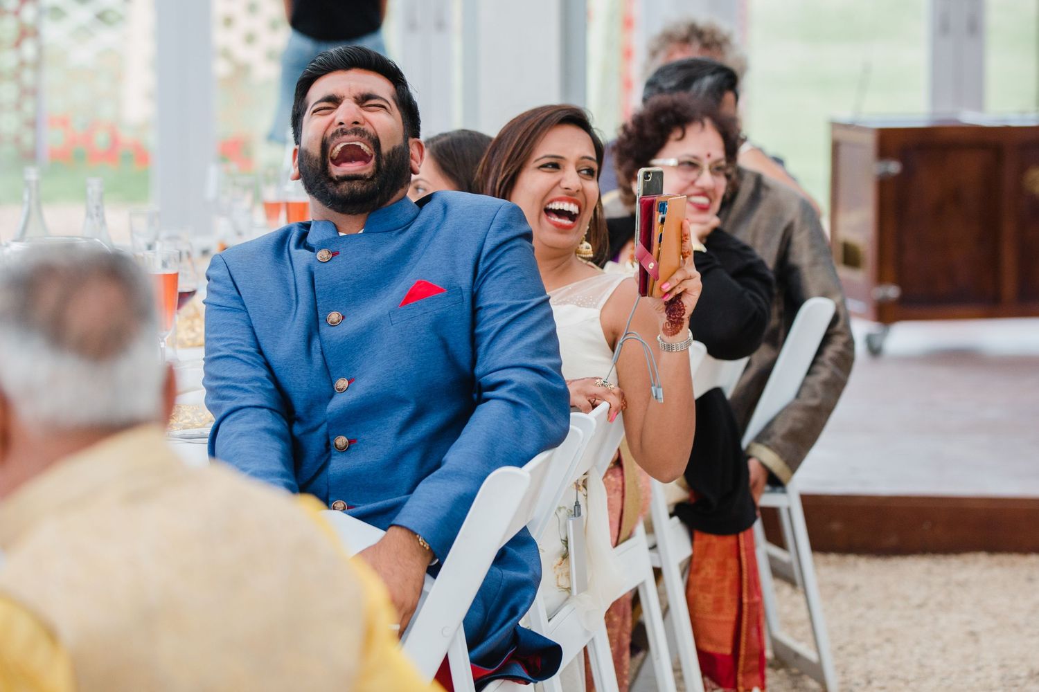 A candid wedding photo of guests at an Indian wedding laughing while someone is making a speech