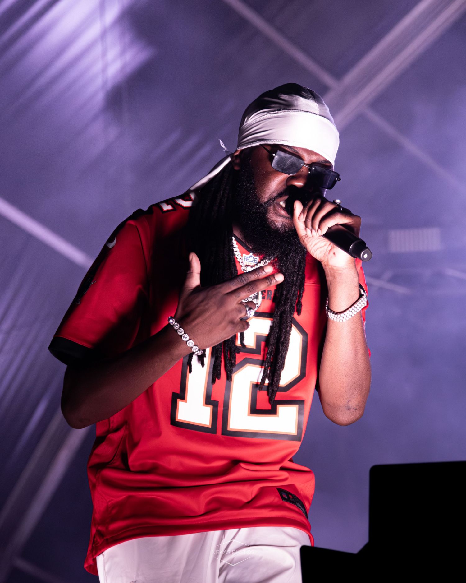 Performer in red jersey number 12 and white headband raps into microphone on stage under dramatic lighting.