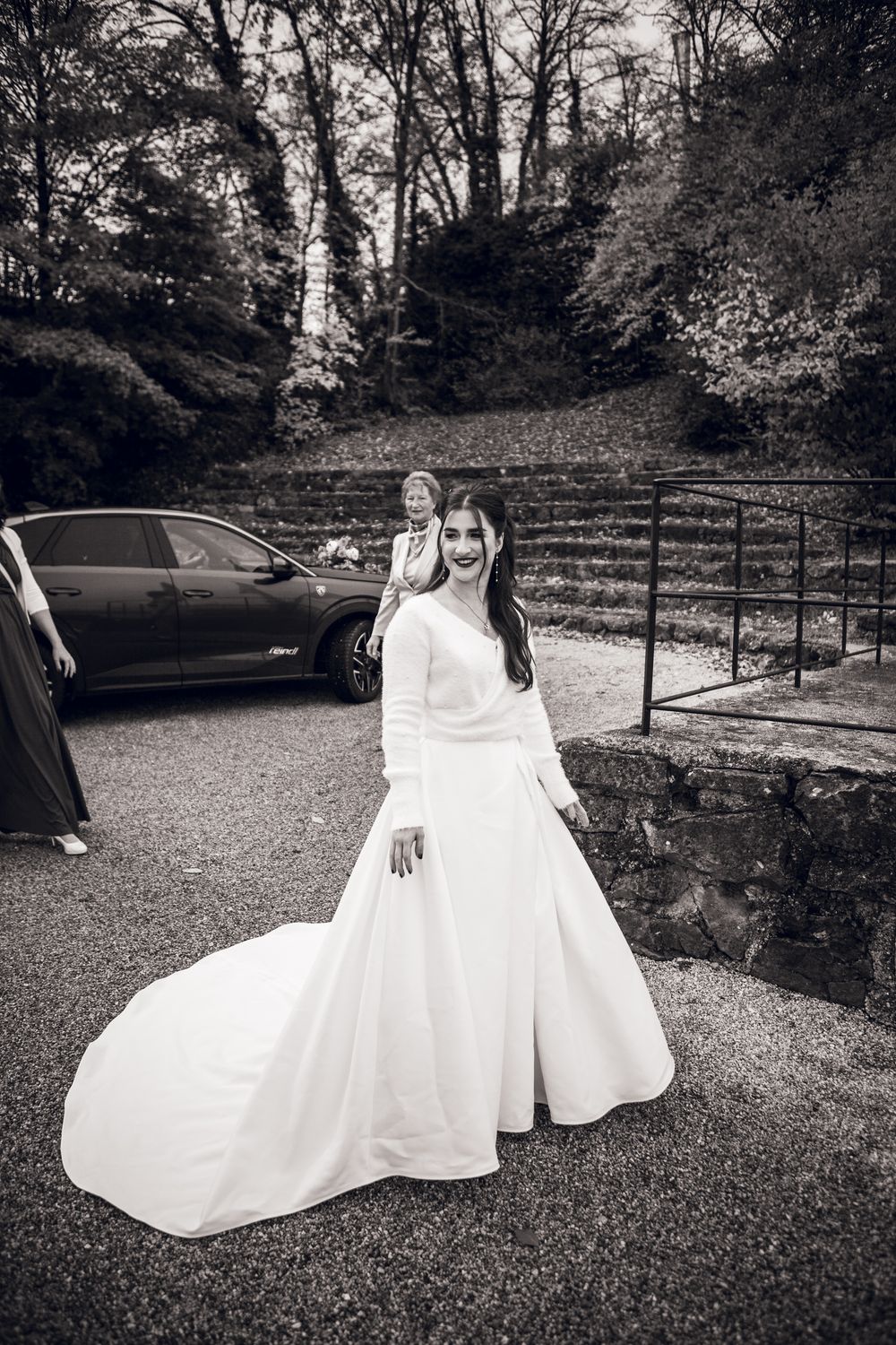 Black and white photo of a smiling bride in a long white gown with train, posing outdoors near stone steps and trees.