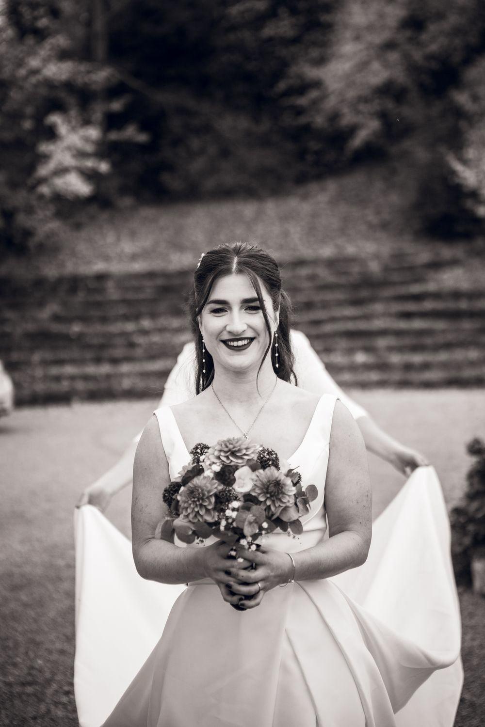 Black and white portrait of a smiling bride in a white gown holding a floral bouquet outdoors.