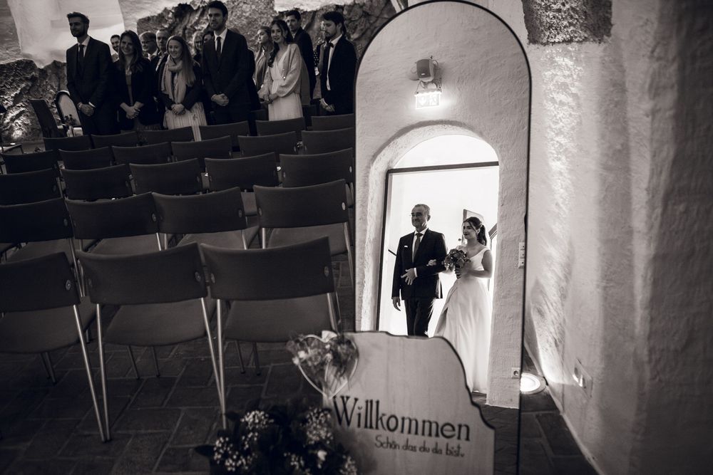 Black and white wedding photo of bride and groom entering a ceremony hall with Willkommen sign.