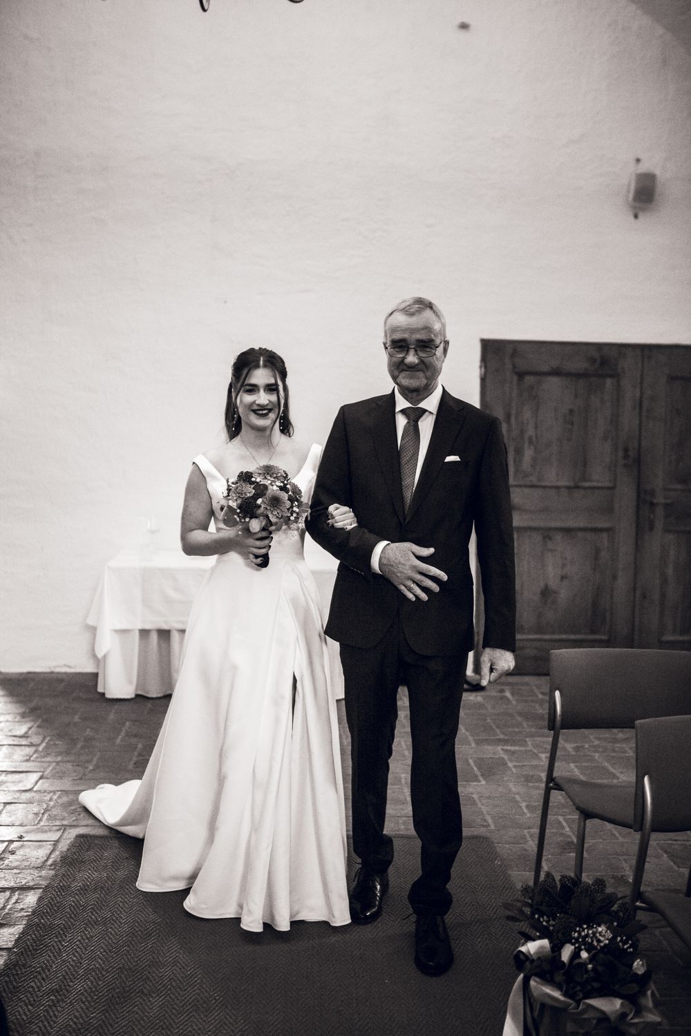 Black and white photo of a bride in a white gown holding a bouquet, walking down the aisle with an older man in a suit.