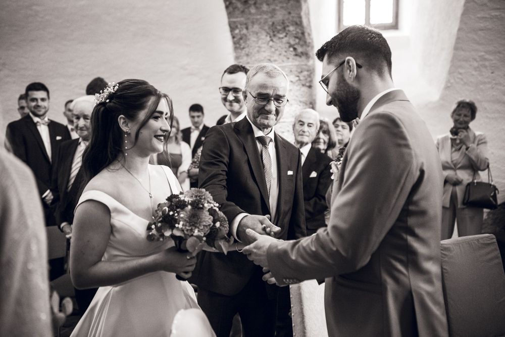 Black and white wedding ceremony photo of bride and groom exchanging rings with officiant and guests in background.