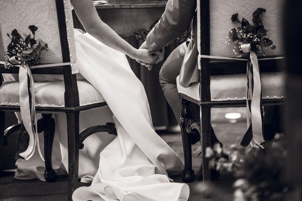 Black and white wedding photo of bride and groom holding hands while seated in decorated chairs during ceremony.