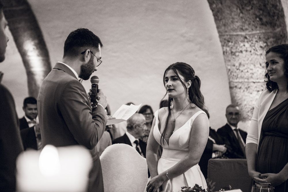 Black and white wedding ceremony photo of bride and groom exchanging vows at an elegant stone venue.