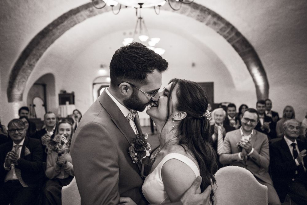 Black and white photo of a couple sharing their first kiss at a wedding ceremony while guests applaud in an arched venue.