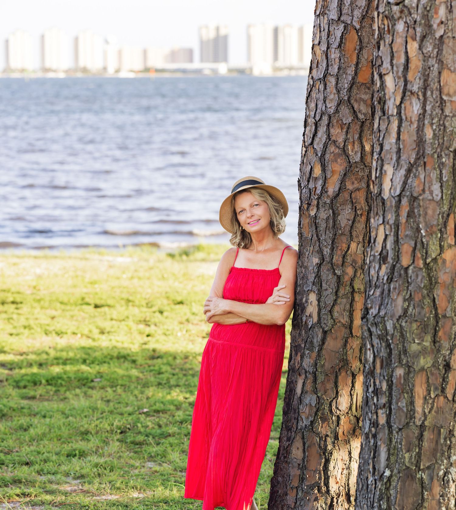 Woman in red dress and straw hat leaning against tree in waterfront park with city skyline in background.