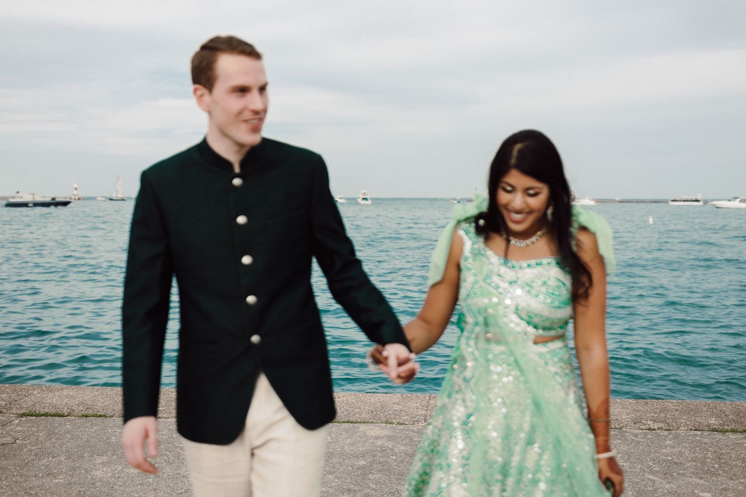 Couple in elegant attire holding hands by the waterfront, man in dark jacket, woman in mint green embroidered lehenga.