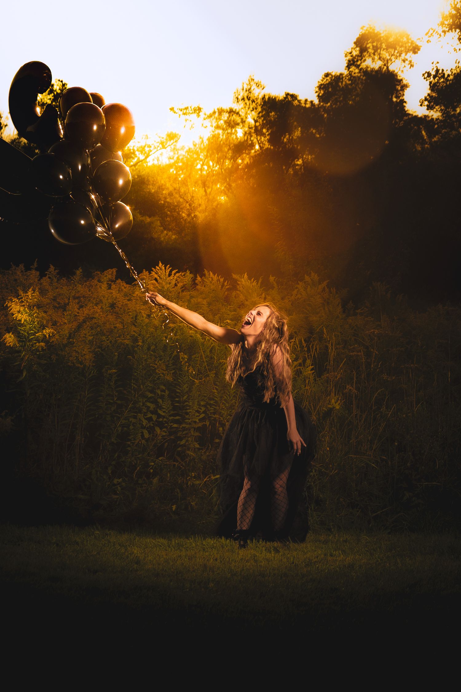 Girl in black dress holding black balloons at sunset in a lush green field with golden light.