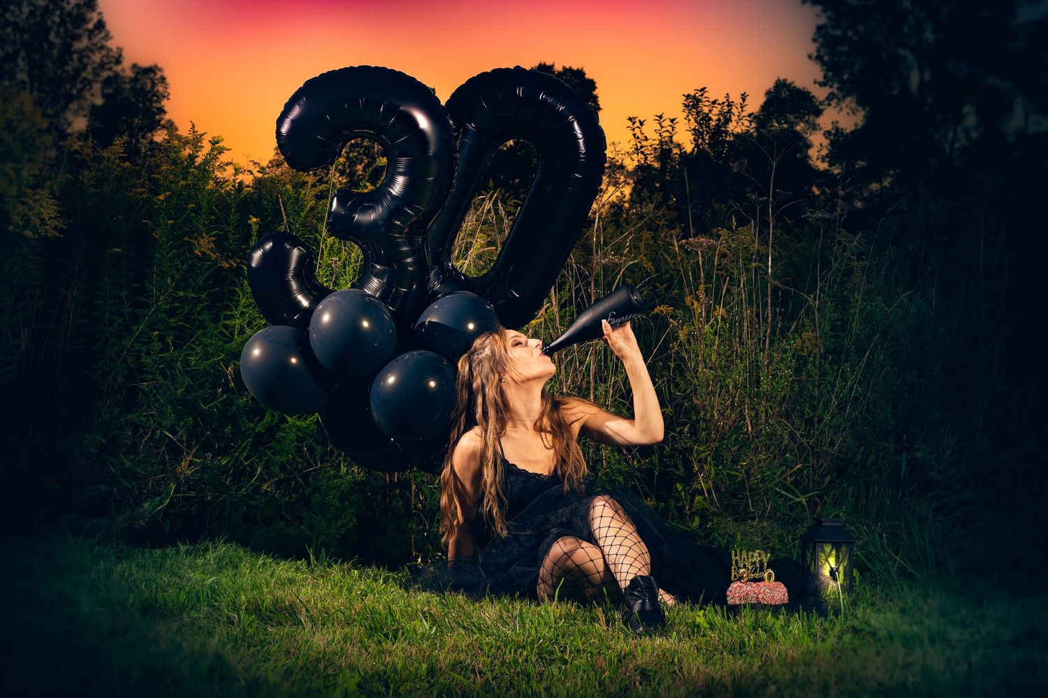 Woman celebrating 39th birthday sitting on grass with black number balloons at sunset.