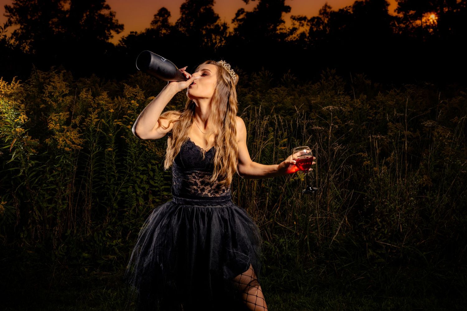 Woman in black dress drinking from bottle at dusk outdoors.