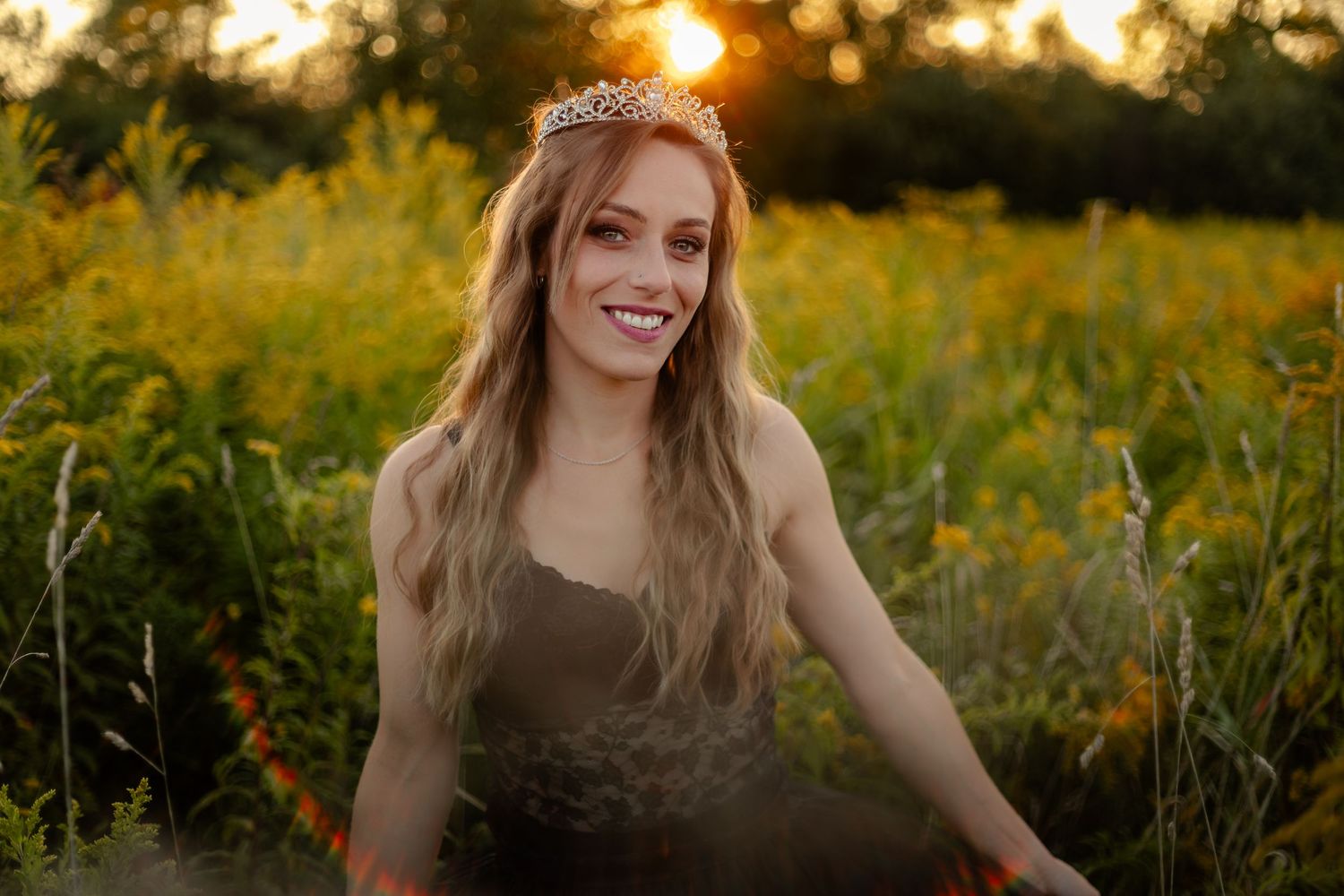 Young woman wearing a tiara smiles in a sunlit field of wildflowers during golden hour.