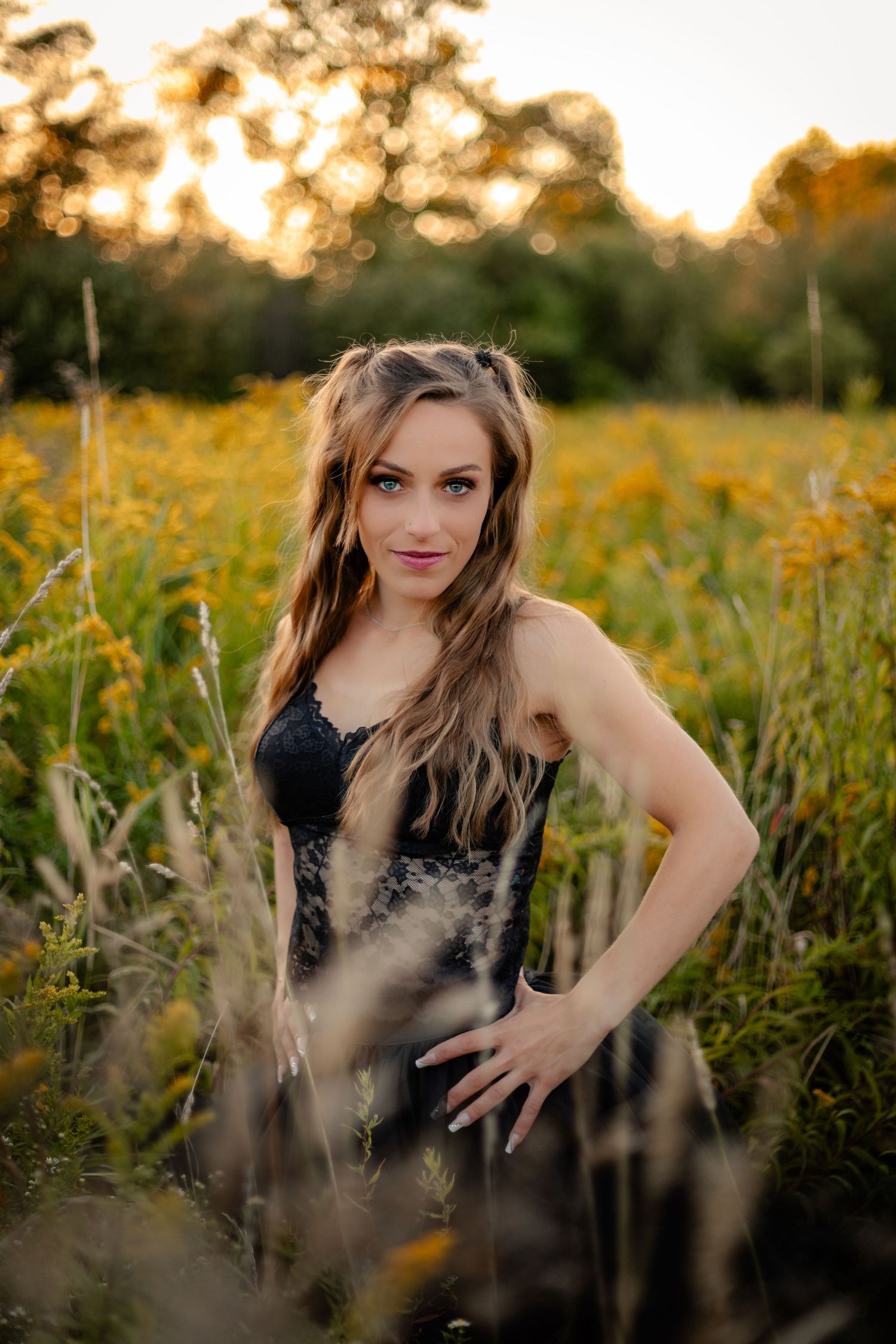 A young woman in a black lace dress poses in a golden wildflower field at sunset.