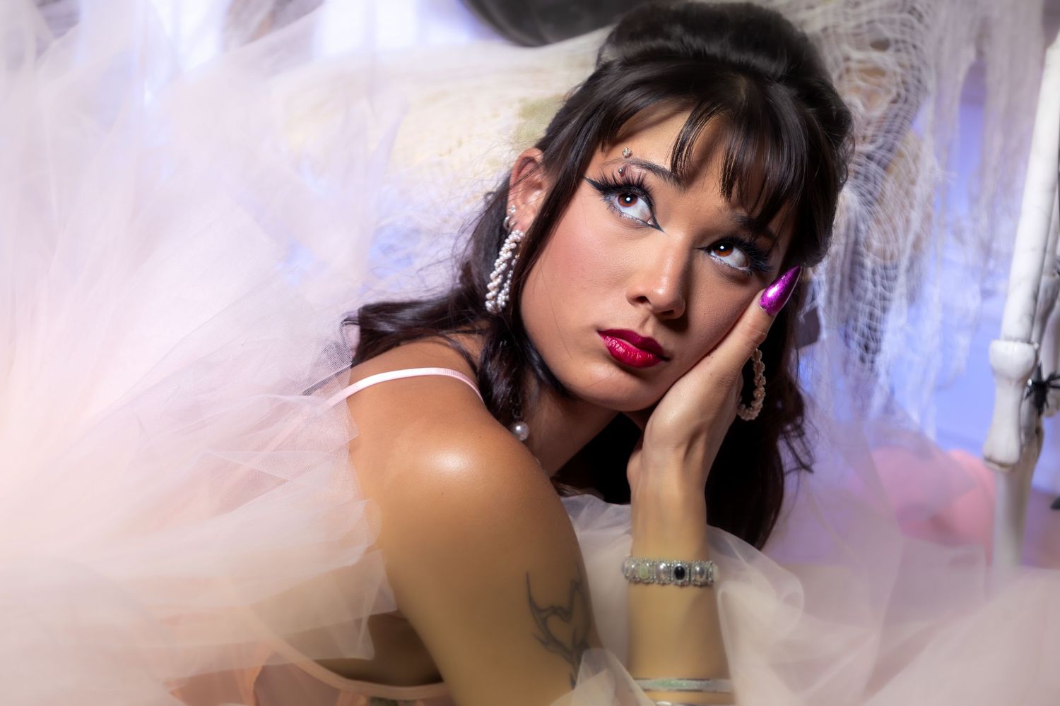 Young woman in white tulle bridal gown with pink nails, diamond earrings, and dark hair with bangs poses thoughtfully.