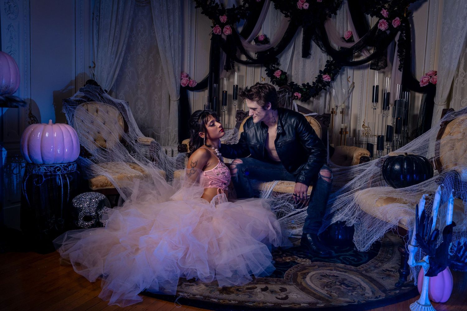 Couple in gothic Halloween wedding attire sitting on ornate furniture surrounded by purple pumpkins and dark decorations.