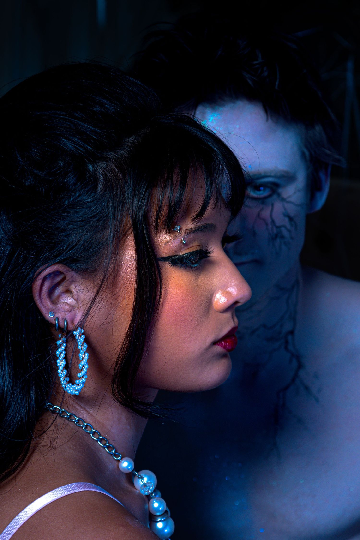 A woman with dramatic makeup and jewelry poses near a blue-lit figure with dark body paint in a moody scene.