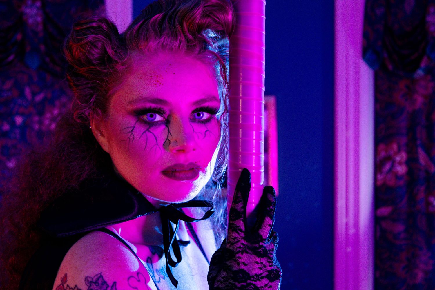 Woman with dramatic gothic makeup, tattoos, lace gloves, and hair buns poses in neon purple and blue lighting.