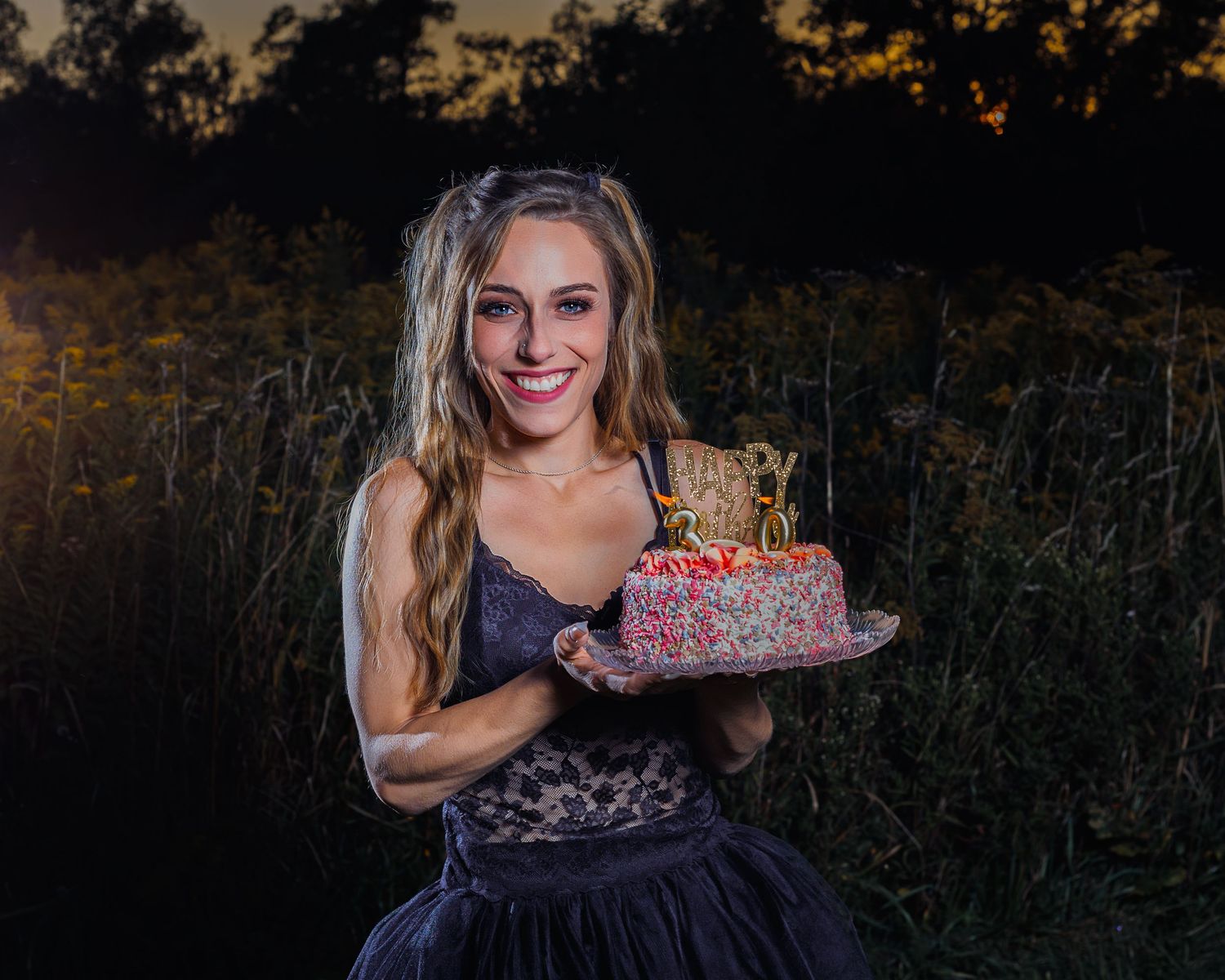 Young woman smiling outdoors at dusk, holding a decorated birthday cake with candles in a field setting.