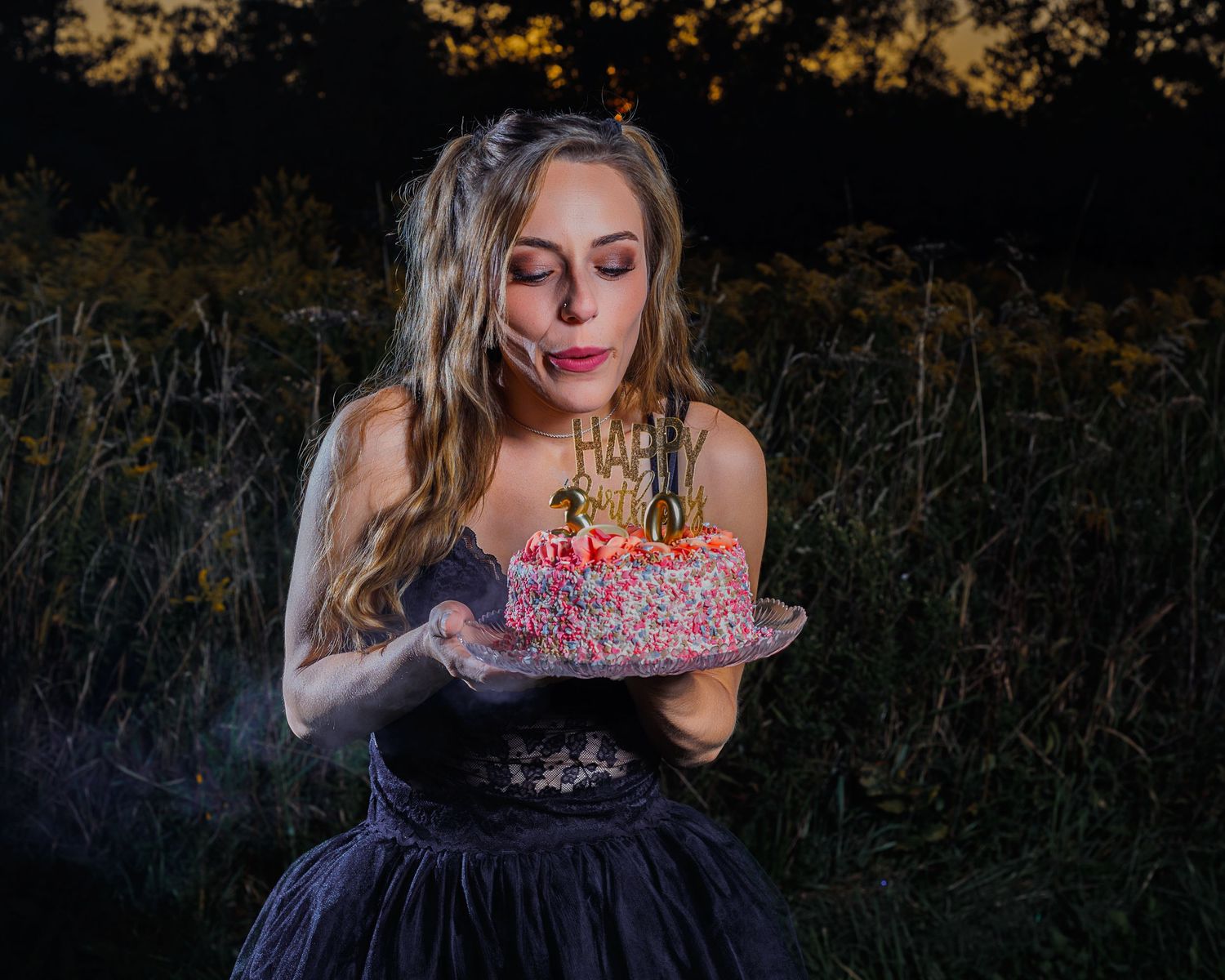 Woman in black dress holding a decorated birthday cake outdoors at dusk near lush greenery.