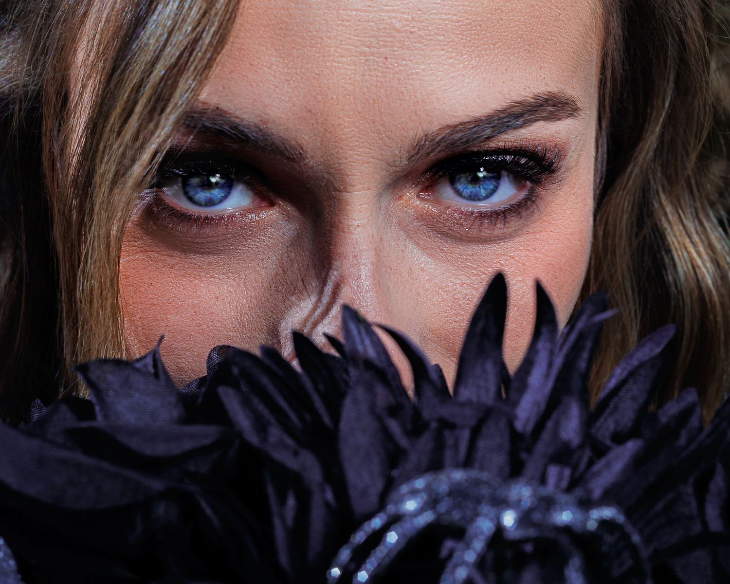 Close-up of a woman with striking blue eyes peering over dark purple feathered flowers with dramatic eye makeup.