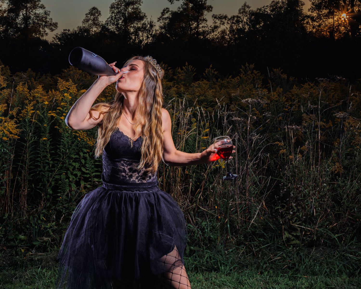 Woman in black dress drinking from a wine bottle while holding a glass in a dramatic outdoor night setting.