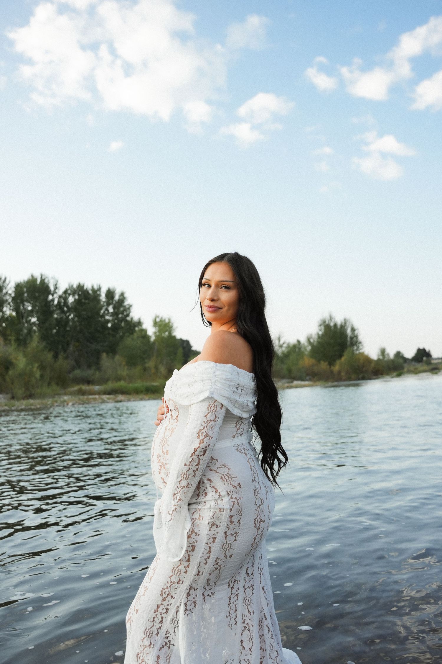 Pregnant woman in white lace off-shoulder gown standing by a serene river with lush green trees and blue sky.