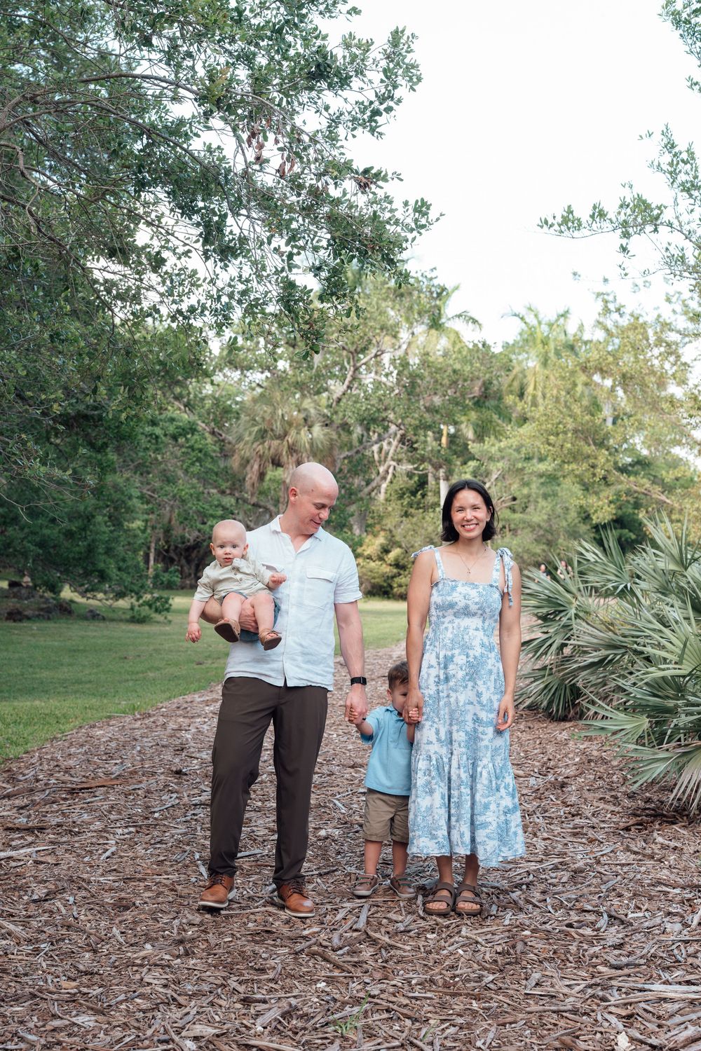 Happy family of four posing on a mulch path in a lush green park, father holding baby and mother holding toddler's hand.
