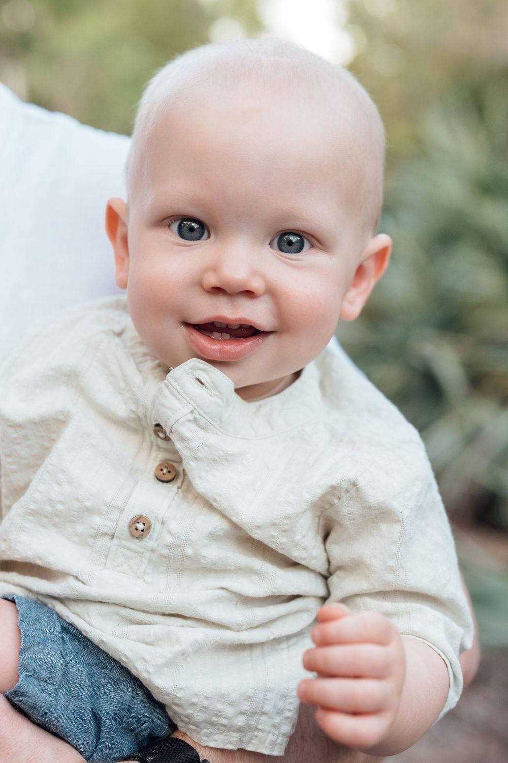 Smiling baby boy with blue eyes wearing a cream linen shirt outdoors.