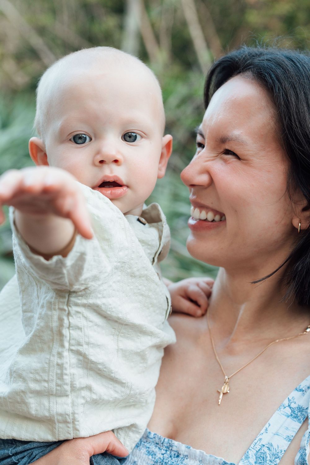 Laughing dark-haired mother holding curious blue-eyed baby boy outdoors.