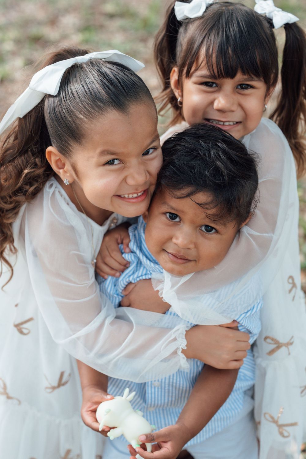 Three young girls in white dresses hug and smile together outdoors.