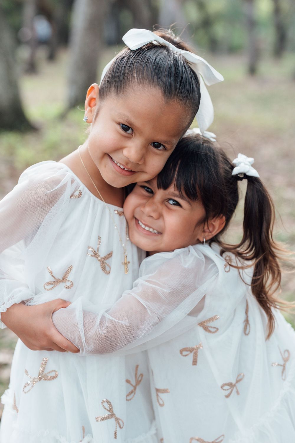 Two young girls in matching white bow-print dresses hug and smile outdoors among trees.