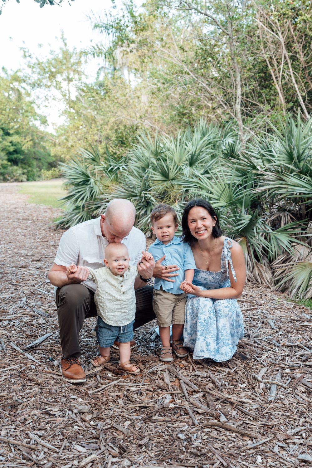 Happy family with two toddlers posing outdoors among tropical palms on a mulch path.