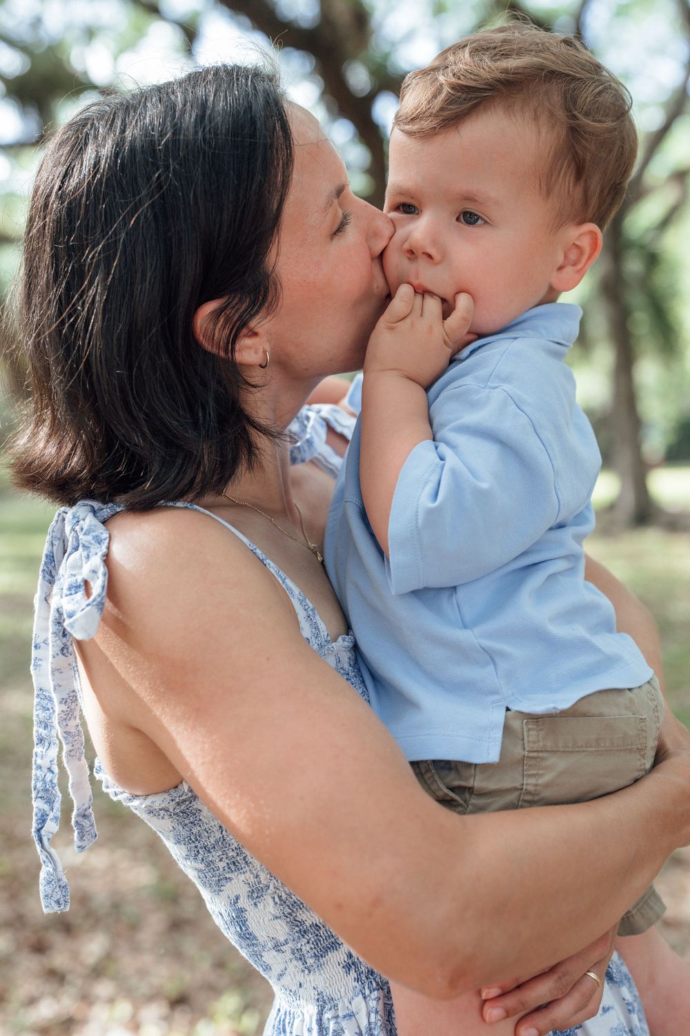 Dark-haired mother in floral dress kissing toddler boy on cheek outdoors.