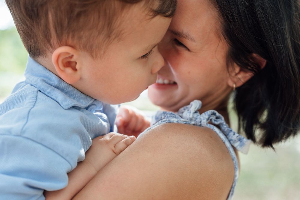 Loving mother holding toddler boy in blue outfit as they touch foreheads and smile at each other.