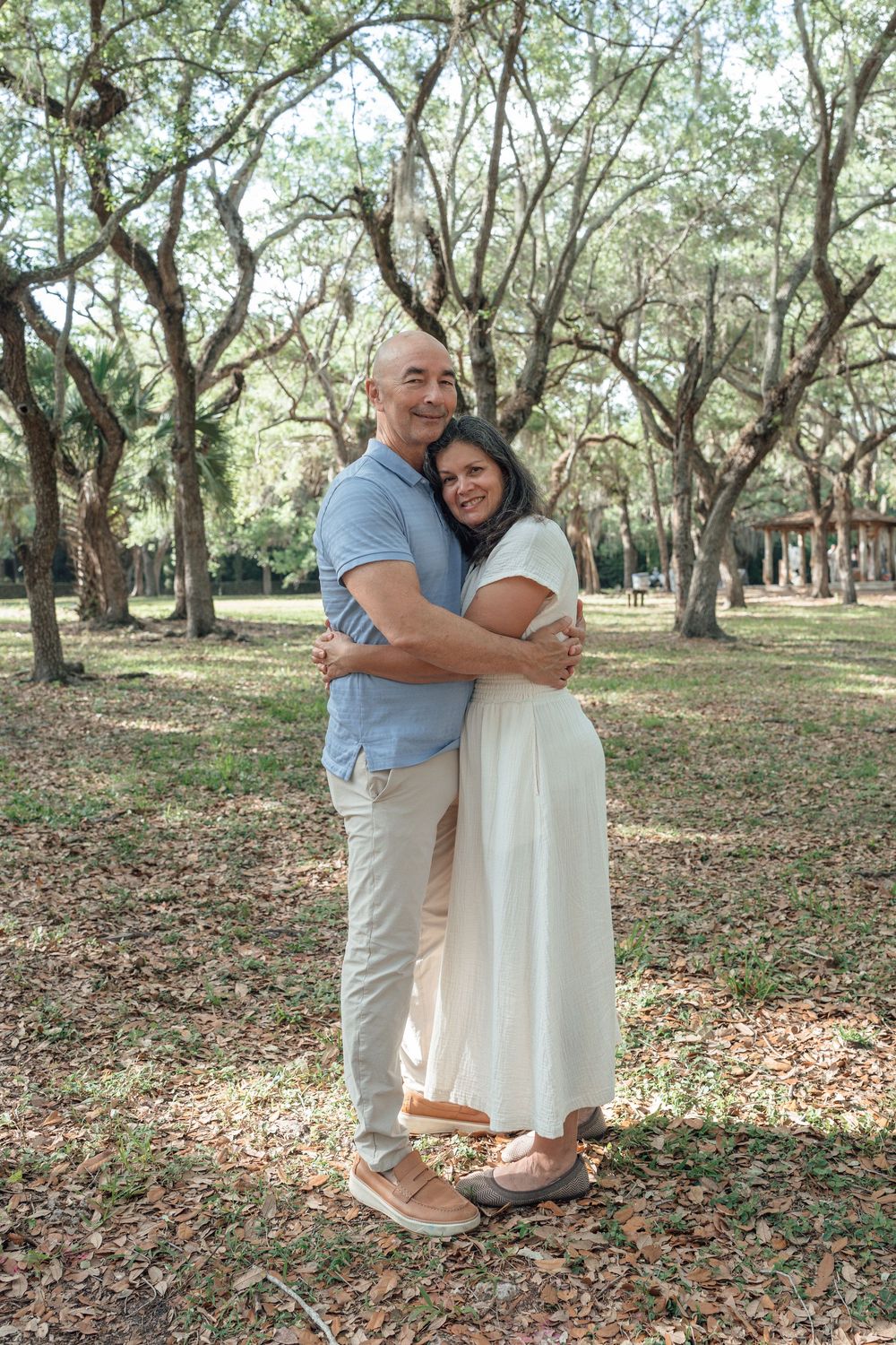 Loving bald man embraces woman in white dress among trees in a park.