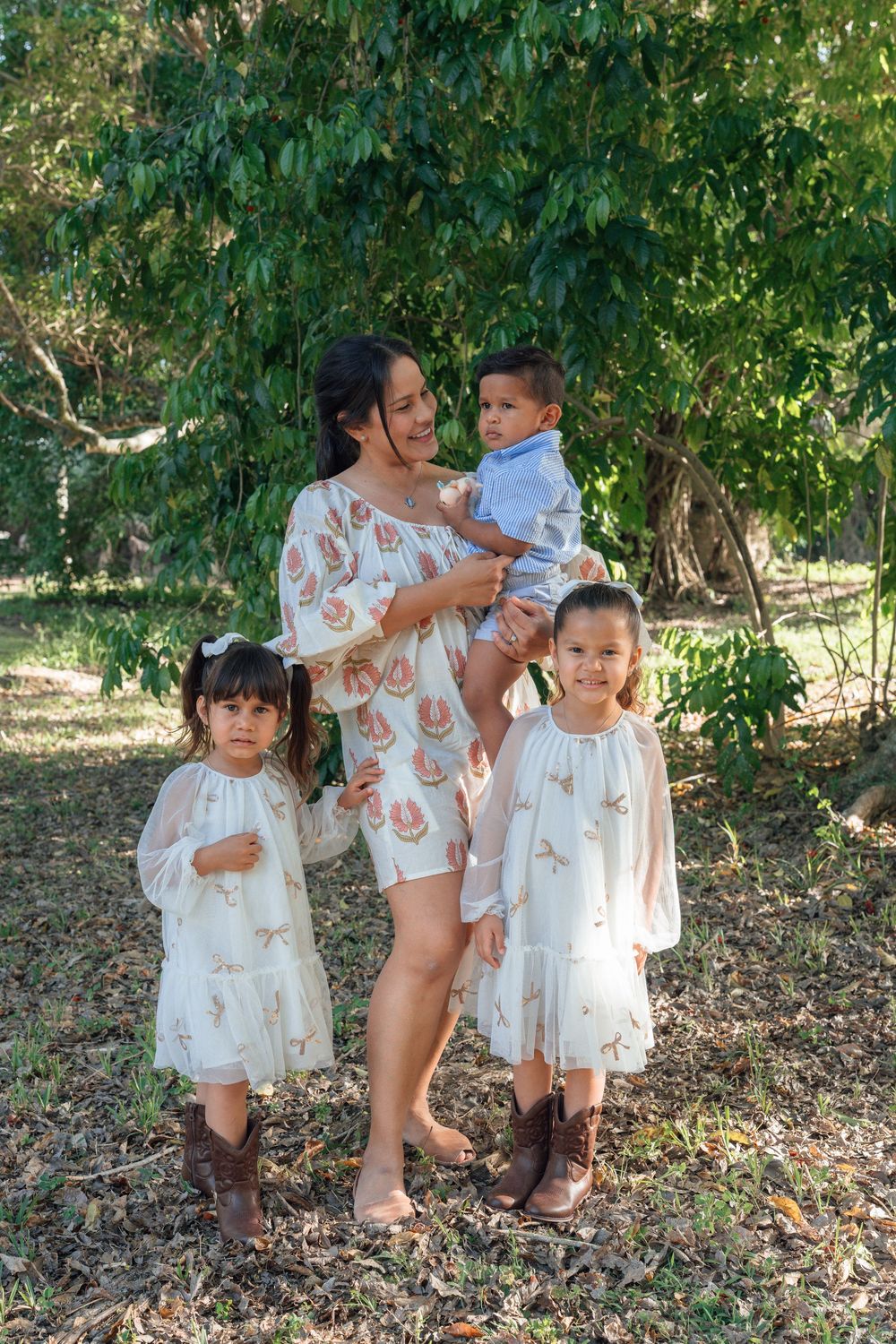 Mother in floral dress holding infant son while posing with two young daughters in white dresses outdoors.
