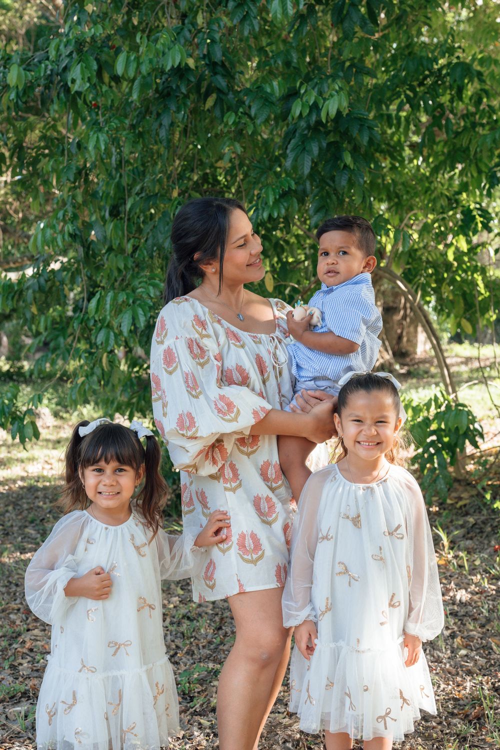 Mother in floral dress holding toddler son while two young daughters smile beside her outdoors.