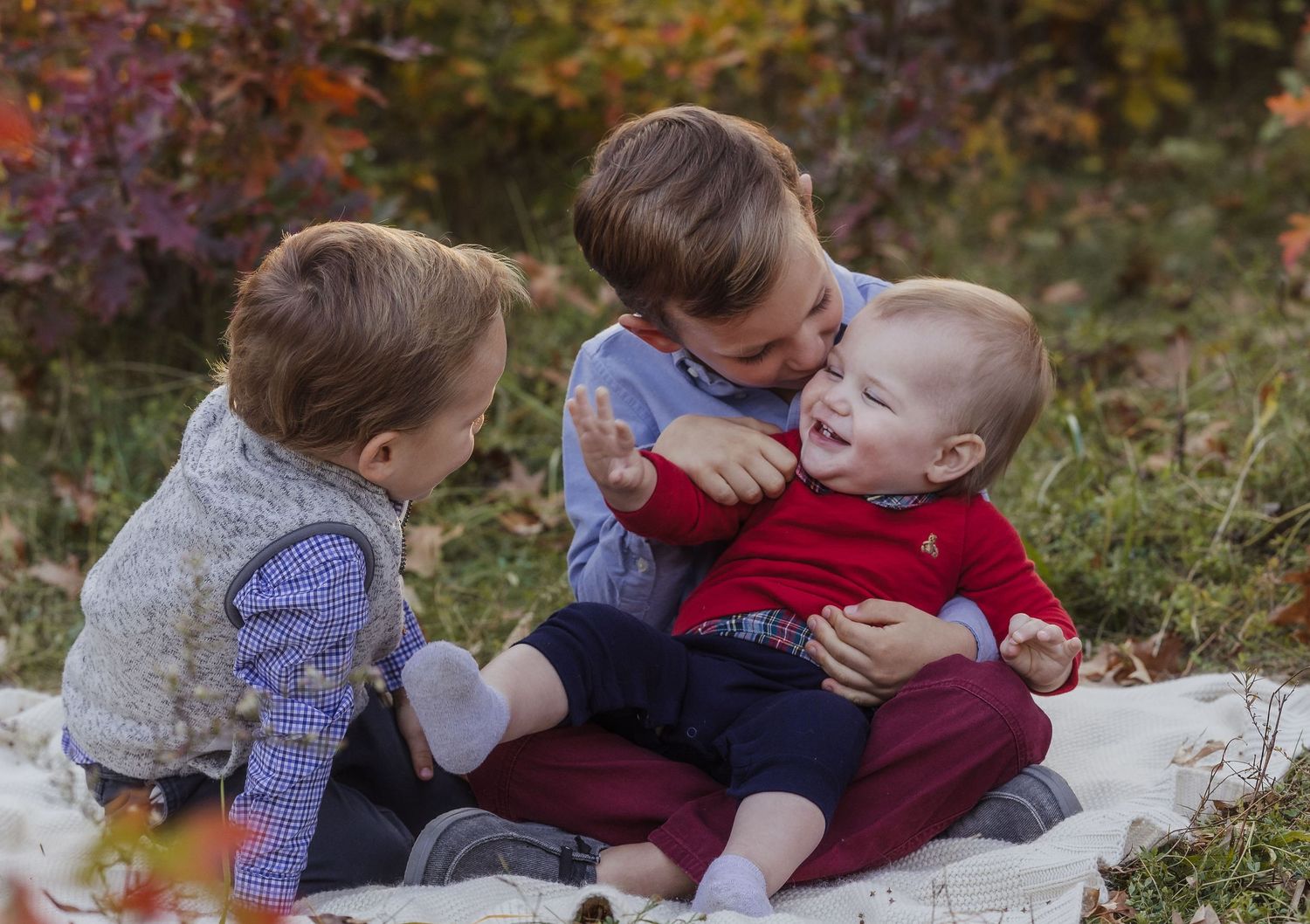 Three young siblings share a joyful moment outdoors in autumn, with the oldest boy kissing the baby while toddler watches.
