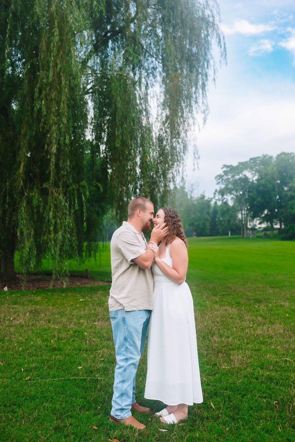 Couple sharing a romantic moment under a weeping willow tree on a lush green lawn.