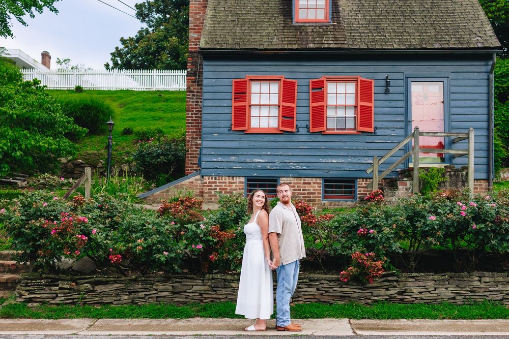 Couple holding hands in front of a charming blue historic home with red shutters and blooming rose bushes.
