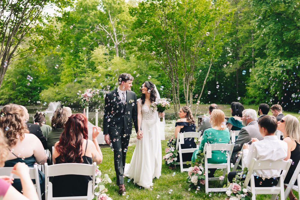 Newlywed couple walks down outdoor aisle amid bubbles, guests seated on white chairs, lush green trees backdrop.