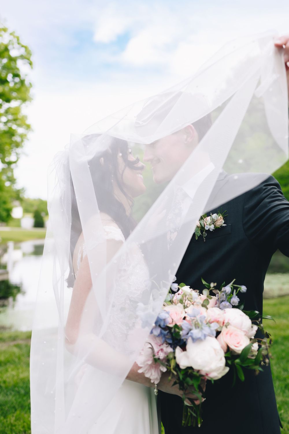 Bride and groom kissing under a flowing white veil, bride holding a pink and blue floral bouquet outdoors.