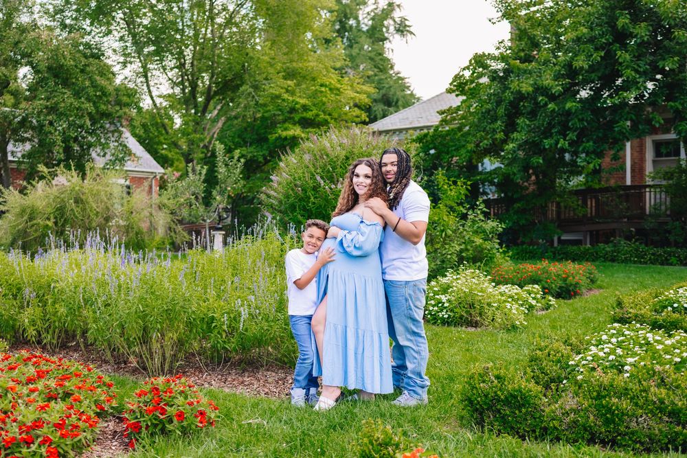 Family of three posing in a lush garden with colorful flowers and green trees in the background.