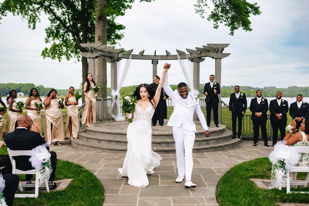 Joyful newlyweds raise hands at outdoor wedding ceremony with bridesmaids in gold and groomsmen in black suits by waterfront pergola.