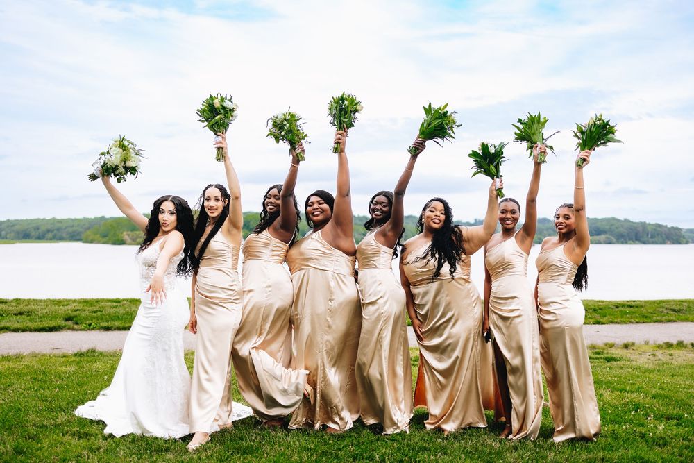 Bride and bridesmaids in gold dresses raising bouquets outdoors by a river on a wedding day.