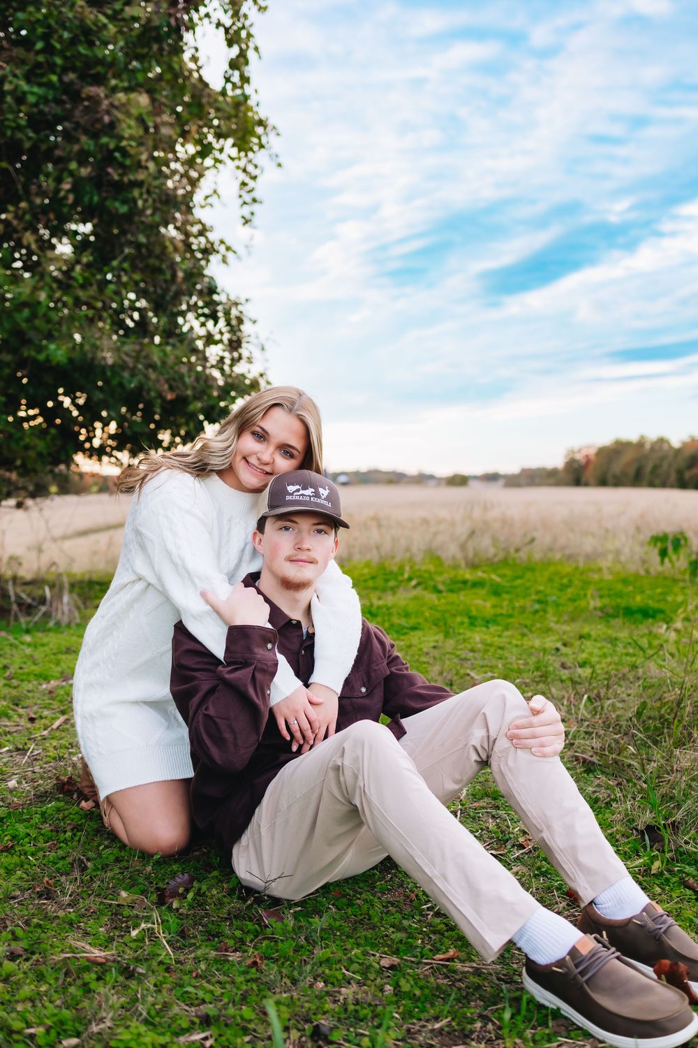 A young woman in a white sweater embraces a boy in a cap sitting on grass with a scenic field backdrop.