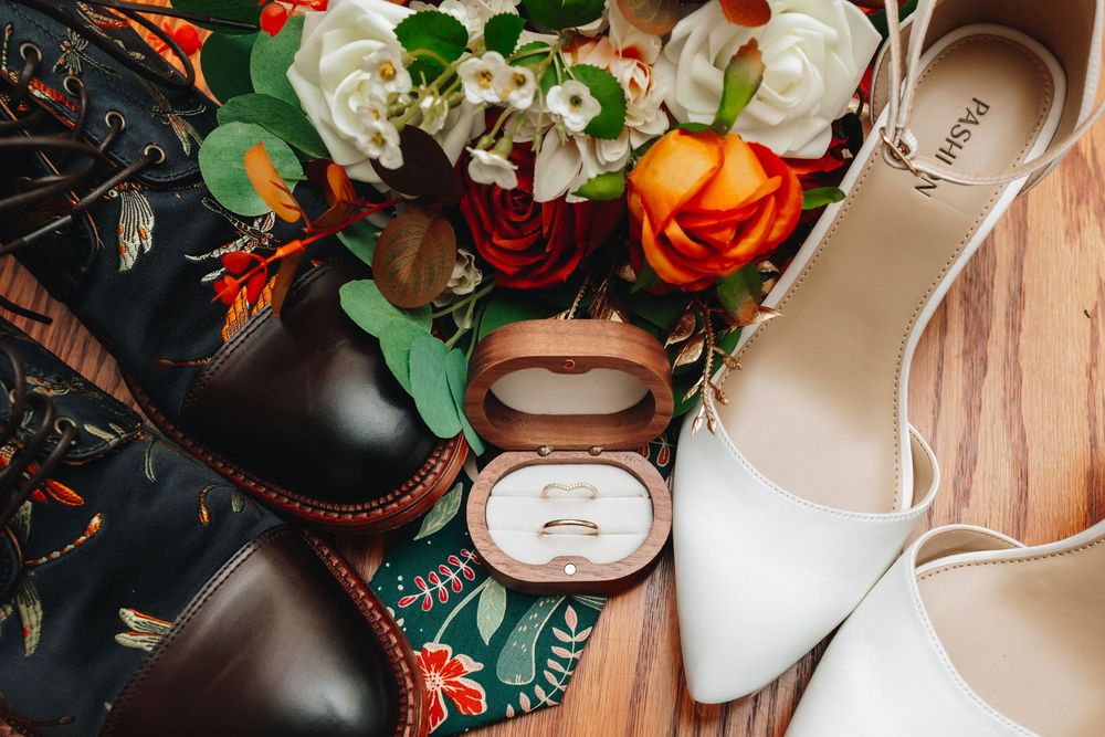 Wedding flat lay with floral bouquet, white heels, brown dress shoes, and wedding rings in a wooden box on wood floor.