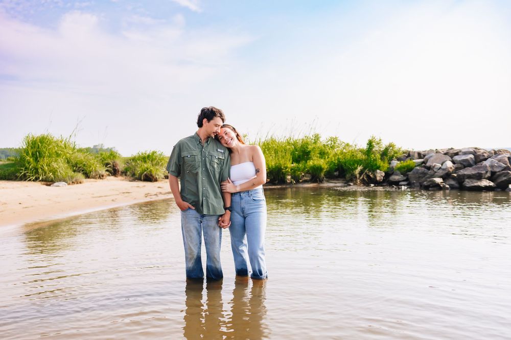 Couple standing in shallow water at a sandy beach shore, embracing romantically with green foliage and rocks behind them.