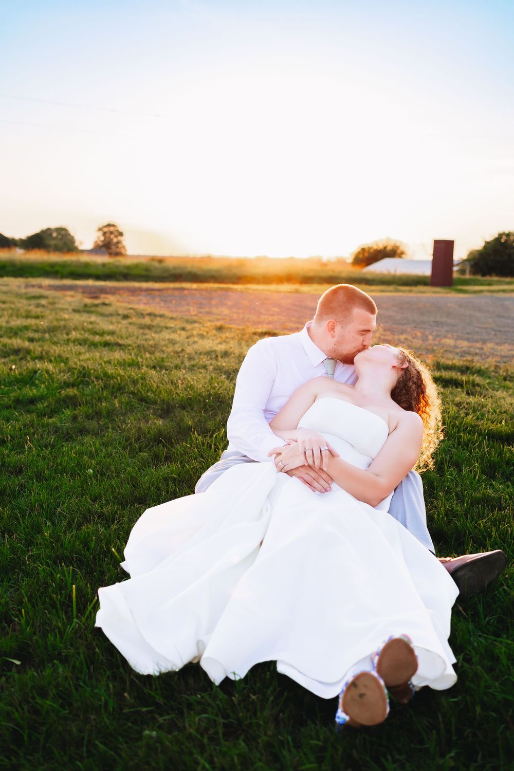 Bride and groom share a romantic sunset kiss on green grass field, bride in white gown cradled in grooms arms.